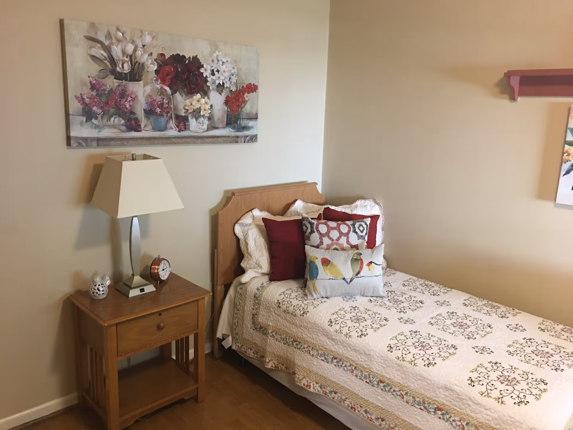 A cozy bedroom with a single bed covered in a white quilt with floral patterns. The bed has several decorative pillows, including one with bird designs. Next to the bed is a wooden nightstand with a lamp, a small clock, and a decorative item. Above the nightstand hangs a painting of various flowers in vases on the wall.