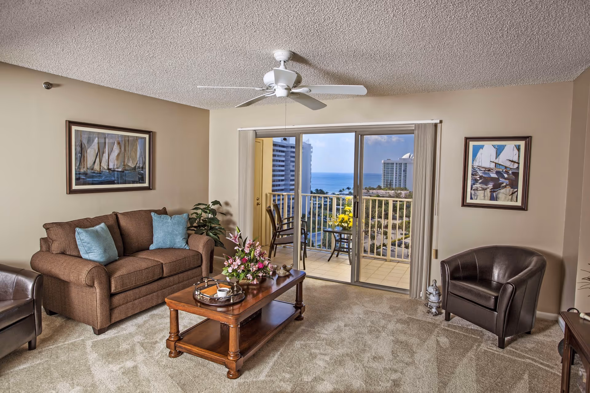 A cozy living room with a brown sofa adorned with two blue cushions, a wooden coffee table with a floral arrangement and a tray, and two dark brown leather chairs. The room has beige walls with framed sailboat artwork and a ceiling fan. Sliding glass doors open to a balcony with a view of tall buildings, greenery, and the ocean in the background.