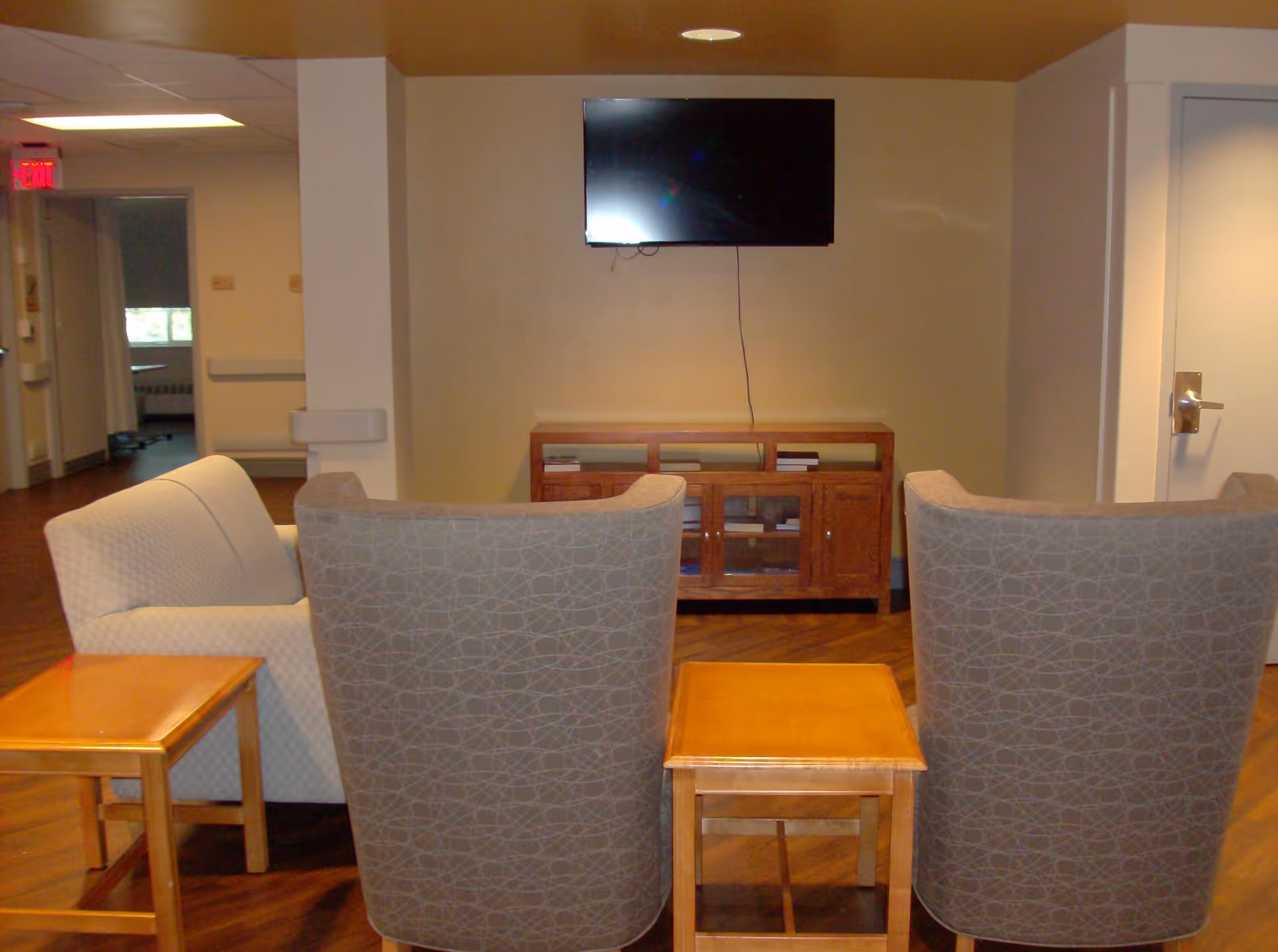 A small seating area with two patterned armchairs and a white couch facing a wall-mounted flat screen TV above a wooden cabinet. There are two wooden side tables between the chairs and couch. The room has wood flooring and beige walls, with a hallway visible to the left and a closed door to the right.