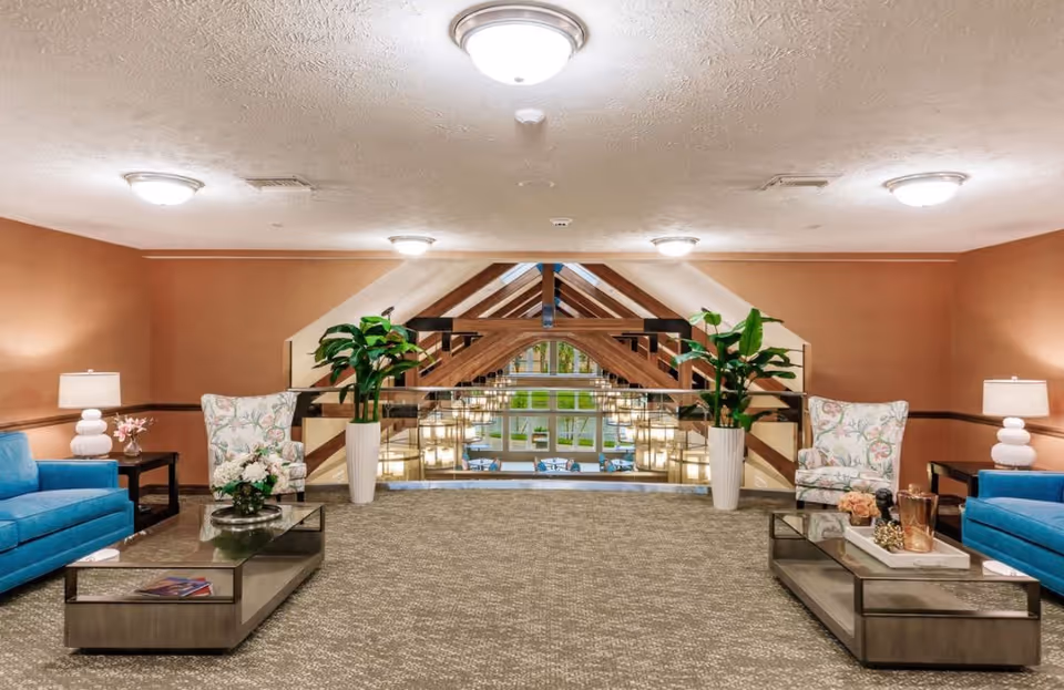 A spacious and well-lit lounge area in a retirement community with two blue sofas, two floral armchairs, two glass coffee tables, potted plants, and table lamps. The area overlooks a large window with wooden beams and hanging lights, showing an outdoor patio with tables and chairs.