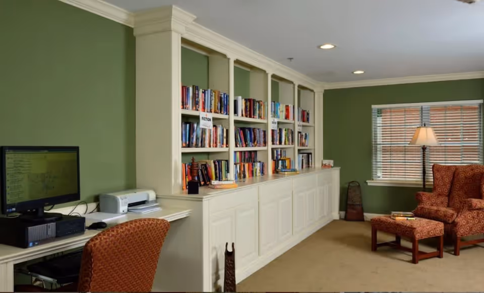 A cozy room with green walls featuring a built-in white bookshelf filled with books labeled 'Fiction'. There is a computer desk with a monitor, printer, and an orange patterned chair on the left side. On the right side, there is a comfortable orange armchair with a matching ottoman, a floor lamp, and a window with blinds.