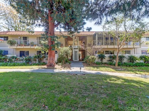 Exterior view of a two-story residential building with balconies, surrounded by trees and greenery, under a clear sky.