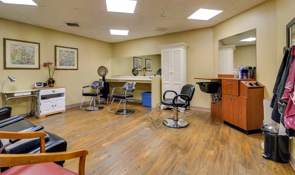 Interior view of a salon area in a senior living facility with three salon chairs, two hair dryer chairs, a wooden cabinet with hair care products, and framed floral artwork on the walls. The room has wood flooring and beige walls with large mirrors.