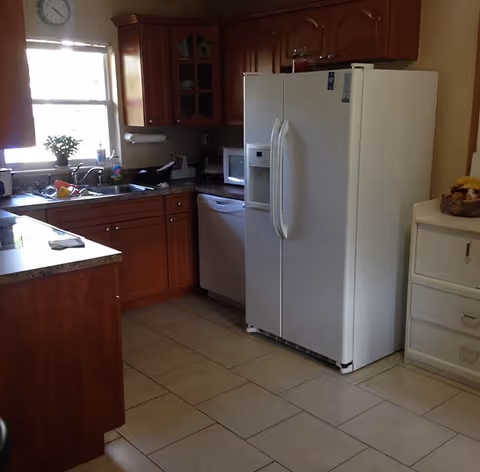 Kitchen with wooden cabinets, a white side-by-side refrigerator, sink under a window, and tiled floor.