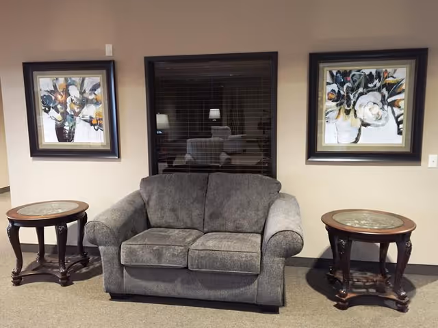 A cozy seating area with a gray upholstered loveseat centered between two round wooden side tables with glass tops. Behind the loveseat is a window with closed blinds reflecting a lamp and chair. On either side of the window are framed floral paintings with dark frames, all set against a beige wall.