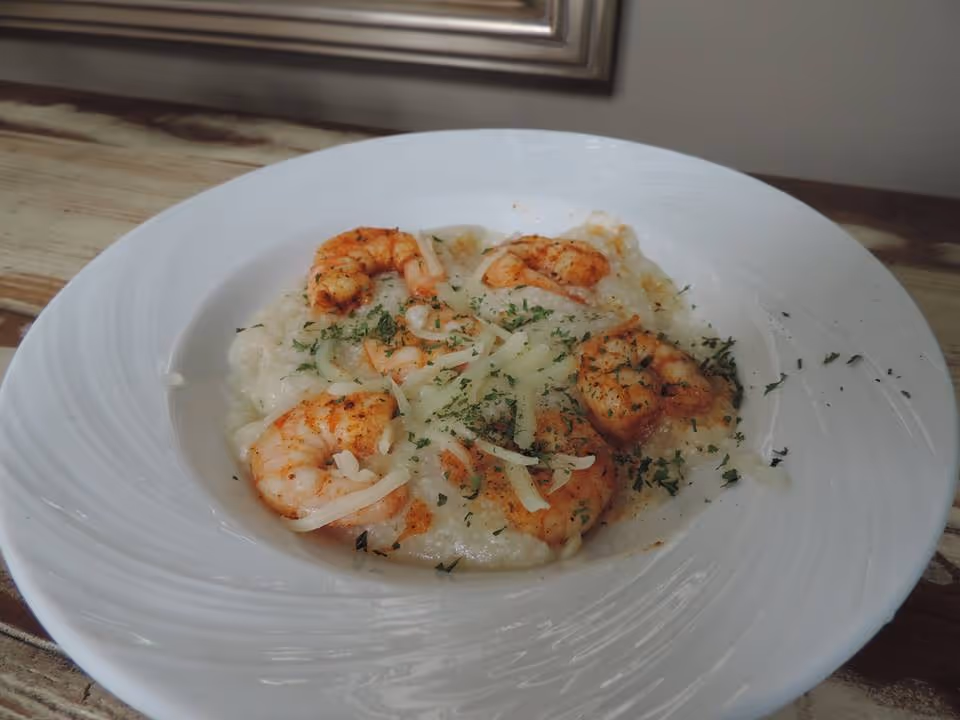 A white plate with shrimp and grits garnished with shredded cheese and herbs, placed on a wooden table near a window.