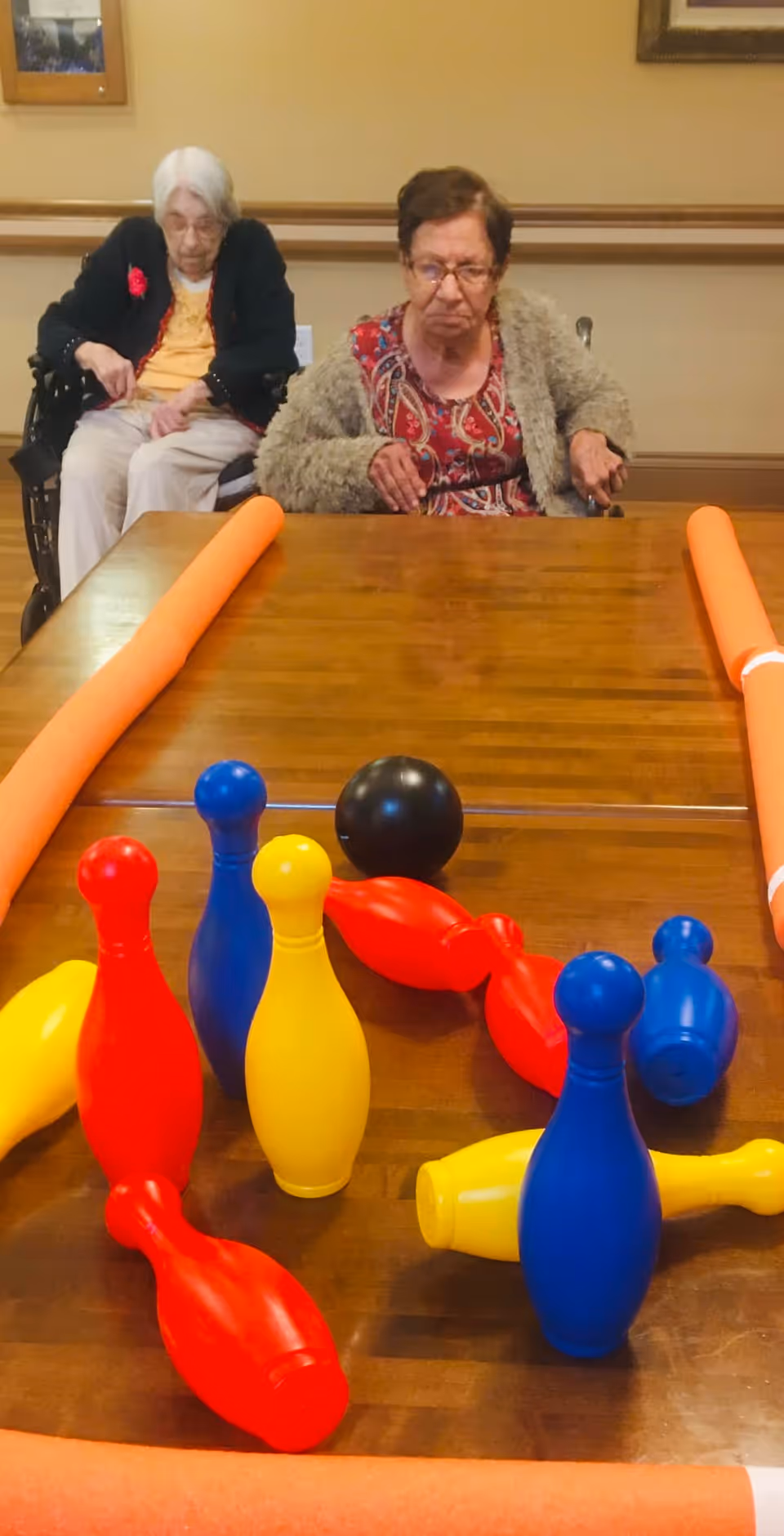 Two elderly women sitting in wheelchairs at a wooden table with colorful plastic bowling pins and balls set up for a game, with orange pool noodles acting as bumpers on the table edges.