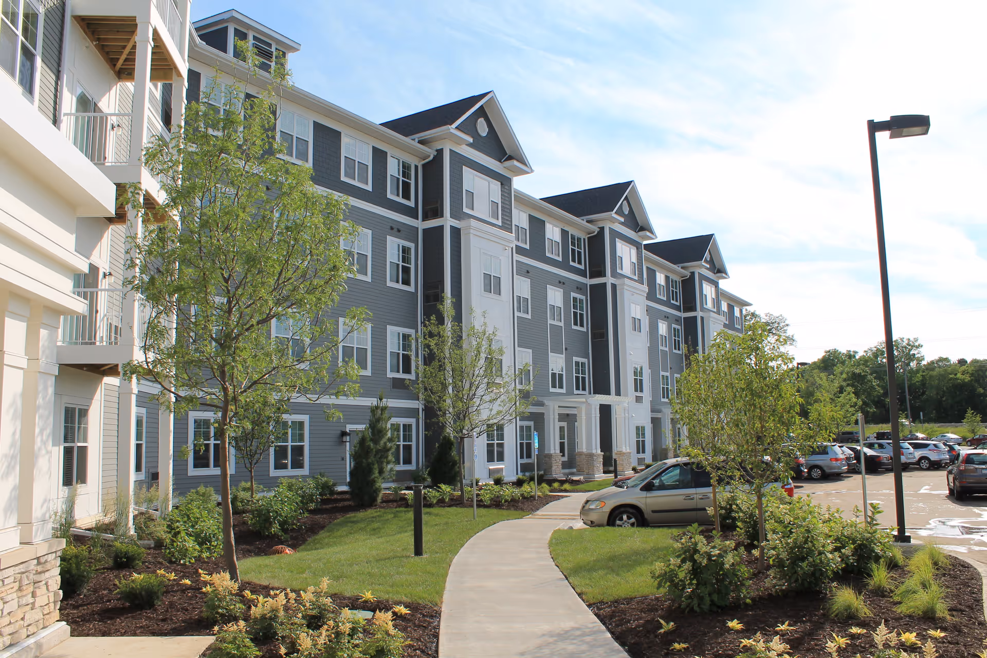 Exterior view of a multi-story senior living facility building with gray siding and white trim. The building is surrounded by landscaped greenery, small trees, and a paved walkway leading to the entrance. Several cars are parked in the adjacent parking lot under a partly cloudy sky.