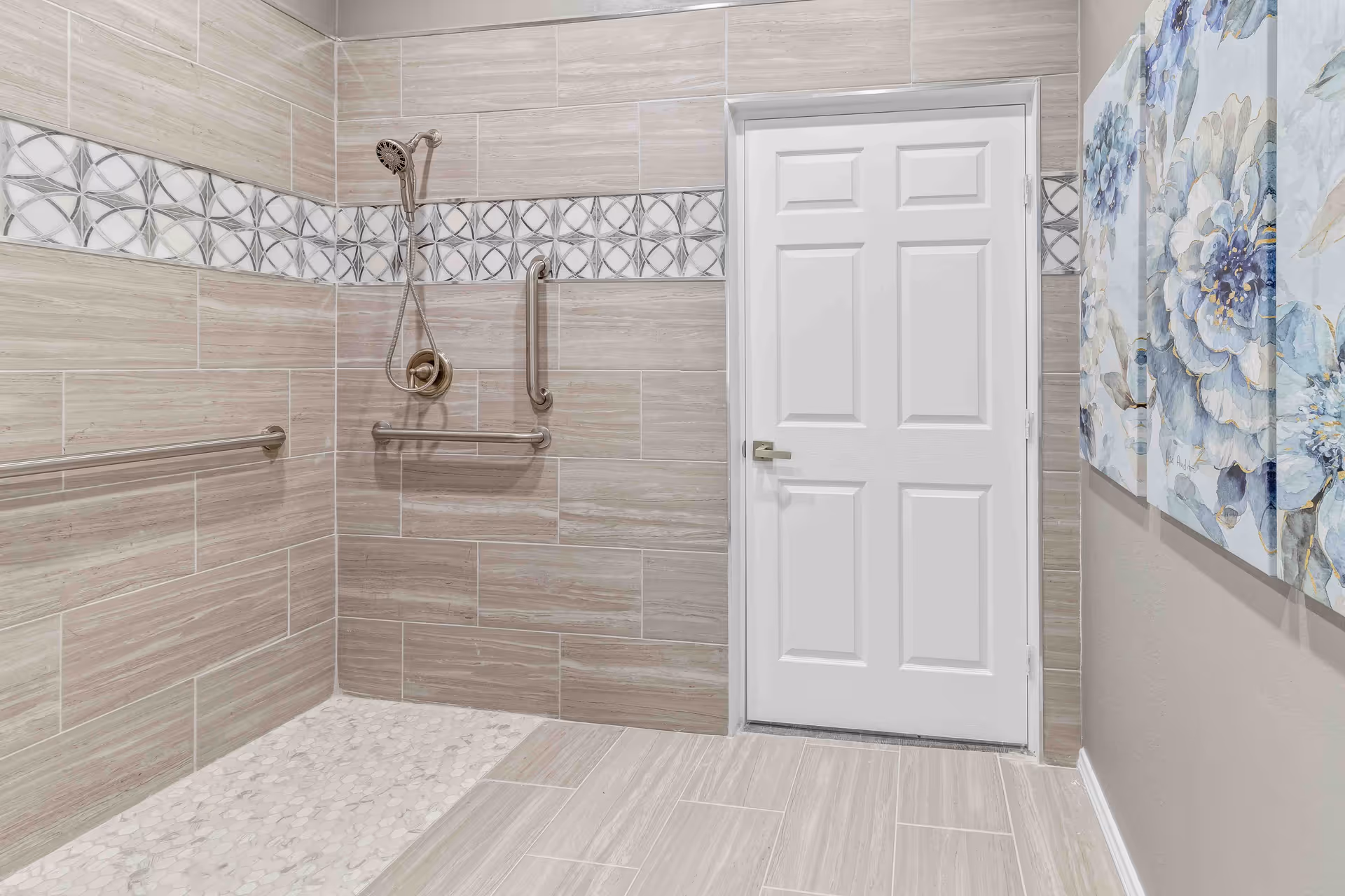 A modern, accessible shower area with beige and light brown tiles, featuring a handheld showerhead, multiple grab bars, and a white door. On the right wall, there are three floral paintings with blue and white flowers.