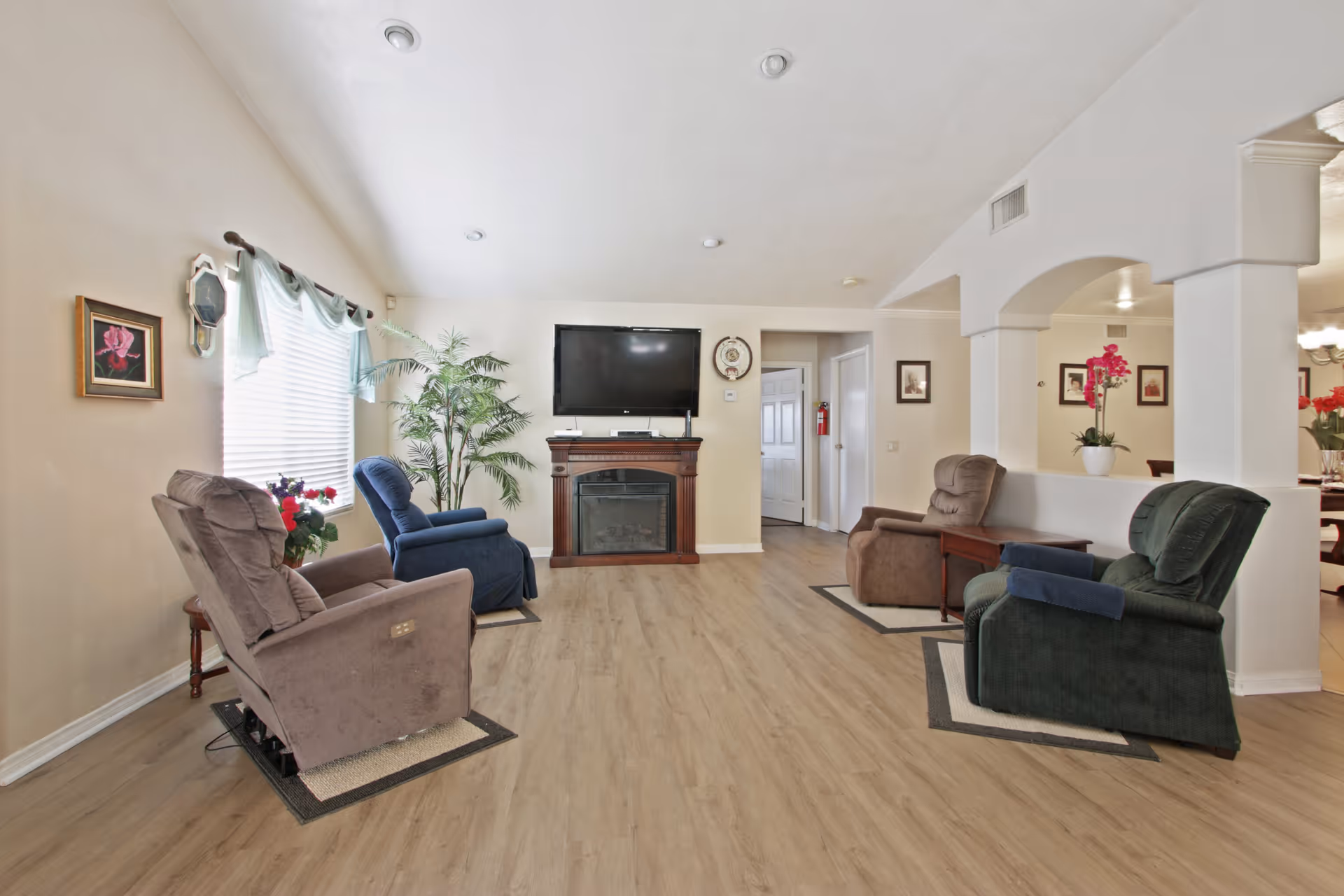 A cozy living room with four recliner chairs arranged around a wall-mounted flat screen TV above an electric fireplace. The room has light-colored walls, wood flooring, a large window with sheer curtains, a potted plant, and some framed pictures on the walls. There is an open archway leading to another room with a dining table and flowers.