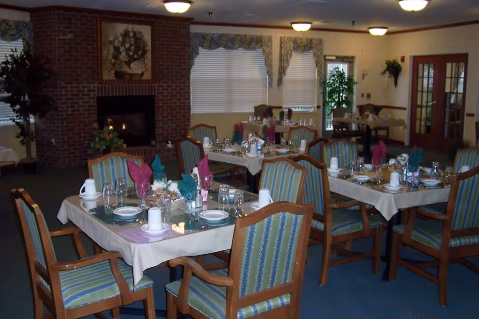 Dining room with multiple tables set for a meal, each table covered with a beige tablecloth and set with plates, glasses, napkins folded in decorative shapes, and utensils. The chairs have wooden frames with green and blue striped upholstery. There is a brick fireplace with a floral painting above it, large windows with valances, and some plants in the room.