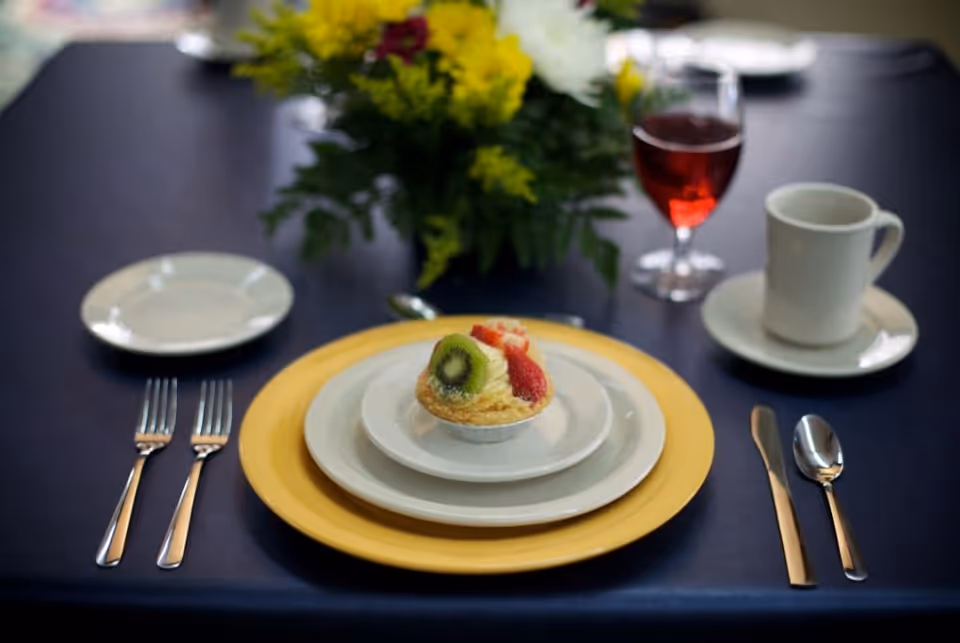 A formal dining table setting with a yellow charger plate, two white plates, and a small tart topped with kiwi and strawberries. The table has a dark blue tablecloth, two forks on the left, a knife and spoon on the right, a white cup and saucer, a glass of red beverage, and a floral centerpiece with yellow and white flowers in the background.