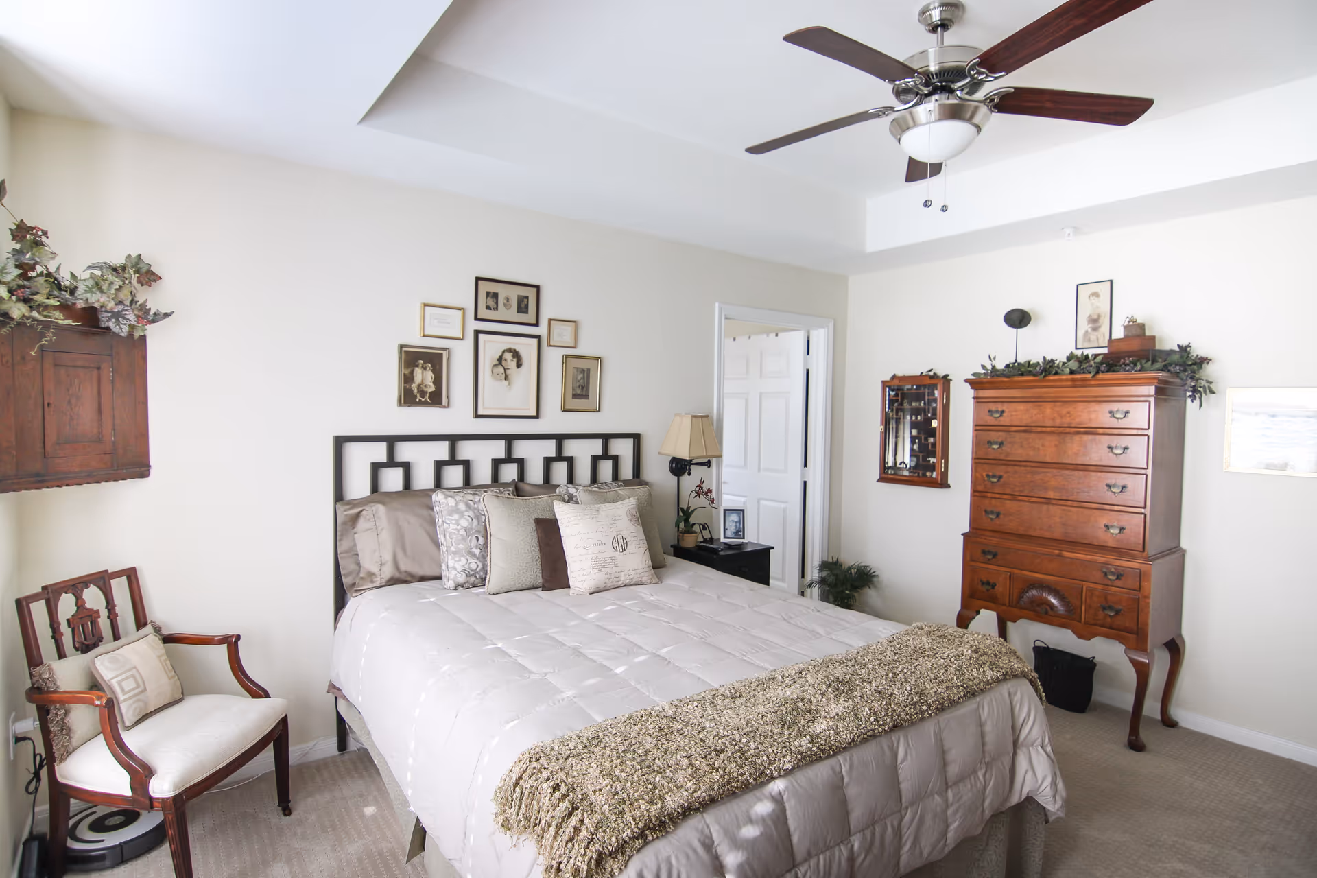 A cozy bedroom with a neatly made bed featuring multiple pillows and a textured throw blanket. The room includes a wooden chair with a cushion, a wooden dresser with decorative greenery on top, framed pictures on the walls, a ceiling fan with dark wooden blades, and a bedside table with a lamp and small plant.