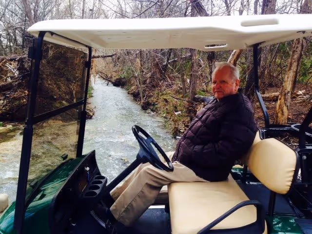 An elderly man wearing a dark jacket and beige pants sits on a beige seat inside a golf cart. The golf cart is parked near a narrow stream surrounded by leafless trees and natural vegetation.