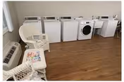 A laundry room with a row of washing machines and dryers along the back wall, wicker chairs and a small table with magazines on a wood floor.