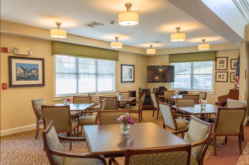 A bright communal dining/activity room with several wooden tables and upholstered chairs, vases of flowers, large windows, and a wall-mounted TV.