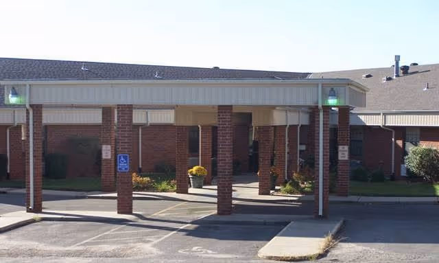 Entrance of a brick building with a covered drop-off area supported by brick columns. There are two green lights on the corners of the roof above the entrance. The area includes a sidewalk and parking spaces, one marked for handicapped parking. Some potted plants and shrubs are visible near the entrance.