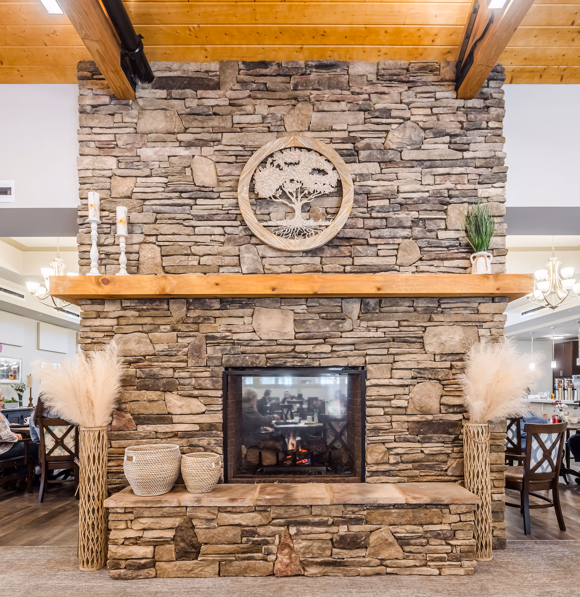 Stone fireplace with a wooden mantel in a senior living facility common area. The mantel is decorated with two white candlesticks on the left and a small plant on the right. On the hearth, there are two woven baskets and two tall vases with pampas grass. Above the mantel, a circular wooden wall art piece depicting a tree is mounted on the stone wall. In the background, dining tables and chairs are visible with people seated.