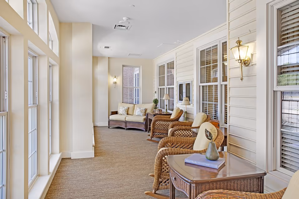 A bright and cozy indoor sitting area with large windows on the left letting in natural light. The space features wicker rocking chairs with cushions, a small table with a decorative vase and a book, and a cushioned loveseat at the far end. Wall-mounted lantern-style lights and table lamps add warmth to the neutral-toned room.