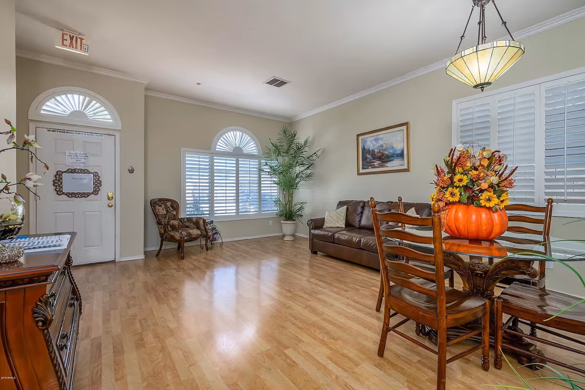 Bright communal living room with hardwood floors, a leather sofa and chairs, a glass dining table topped with a pumpkin floral centerpiece, and a front door with an EXIT sign.