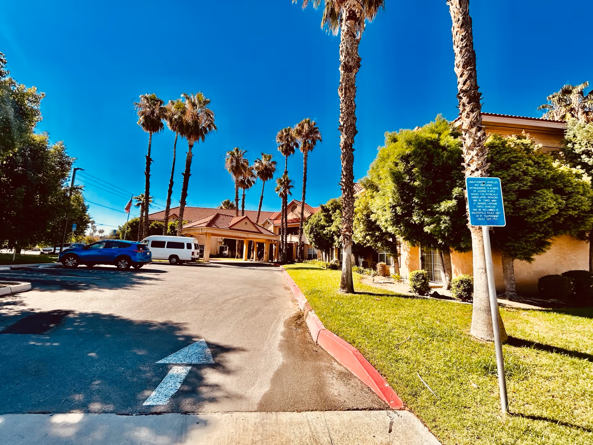 Exterior view of Hallmark Of Bakersfield facility with a driveway leading to the entrance, surrounded by palm trees and green bushes under a clear blue sky. Several parked vehicles are visible near the entrance.