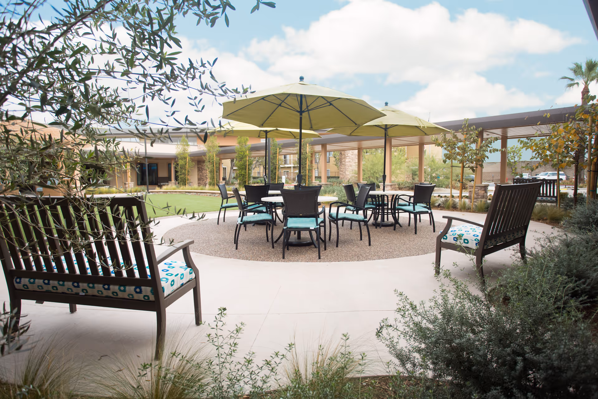 Outdoor seating area at Emerald Court with round tables and chairs under large green umbrellas, surrounded by benches with patterned cushions and landscaped greenery under a partly cloudy sky.