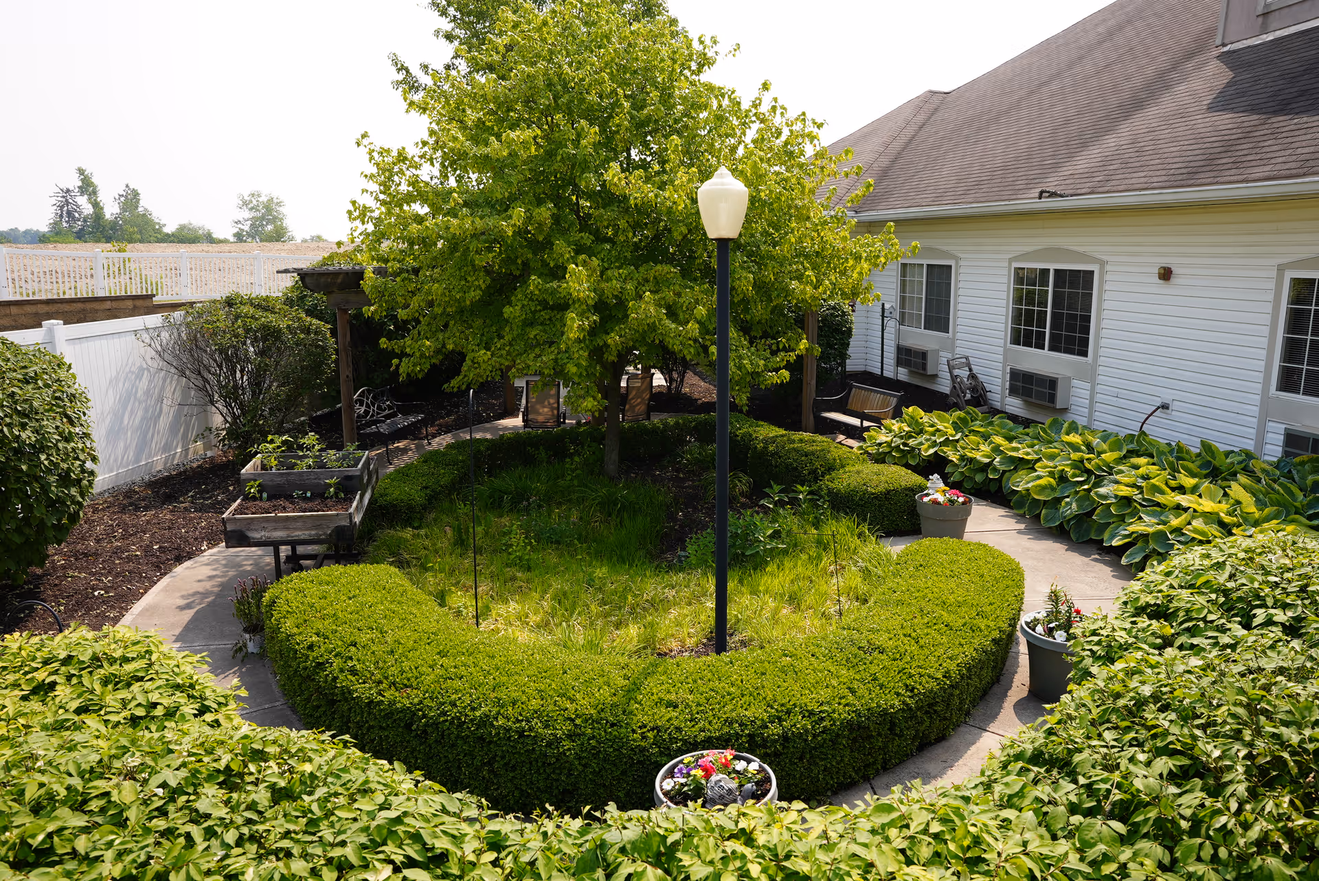 A well-maintained outdoor garden area at Coventry Meadows featuring a circular hedge surrounding a tree with a lamp post in the center. The garden is bordered by a white fence and adjacent to a building with white siding and multiple windows. There are potted plants and benches along the pathway.
