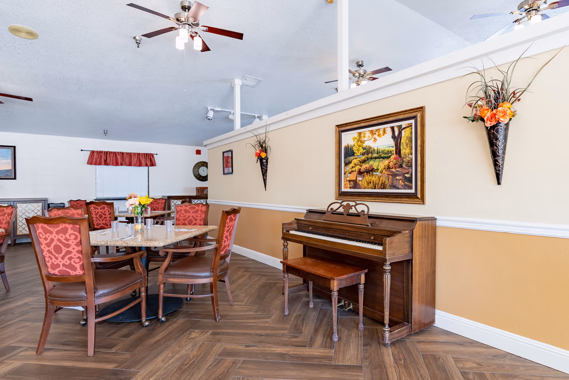 Bright communal dining area with a round table and red upholstered chairs and an upright piano against a half wall decorated with a framed painting and wall planters.