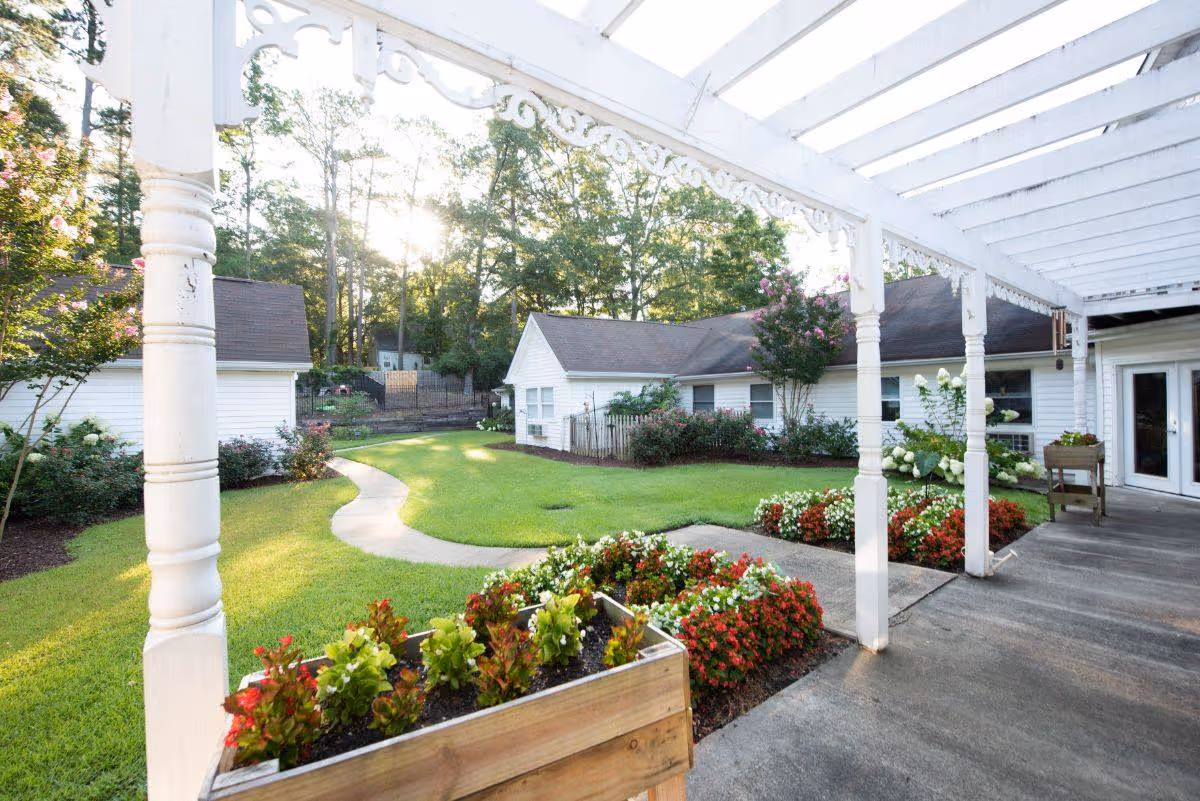 A bright outdoor garden area at Highlands Senior Living Norcross featuring a white pergola with decorative trim, a concrete walkway winding through a well-maintained lawn, colorful flower beds, and white buildings surrounded by trees in the background.