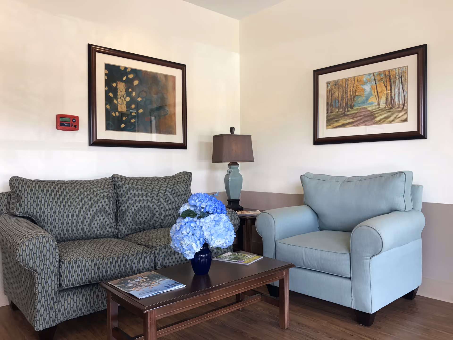 A cozy sitting area with a patterned sofa, light blue armchair, wooden coffee table holding a vase of blue flowers, and framed artwork on the walls.