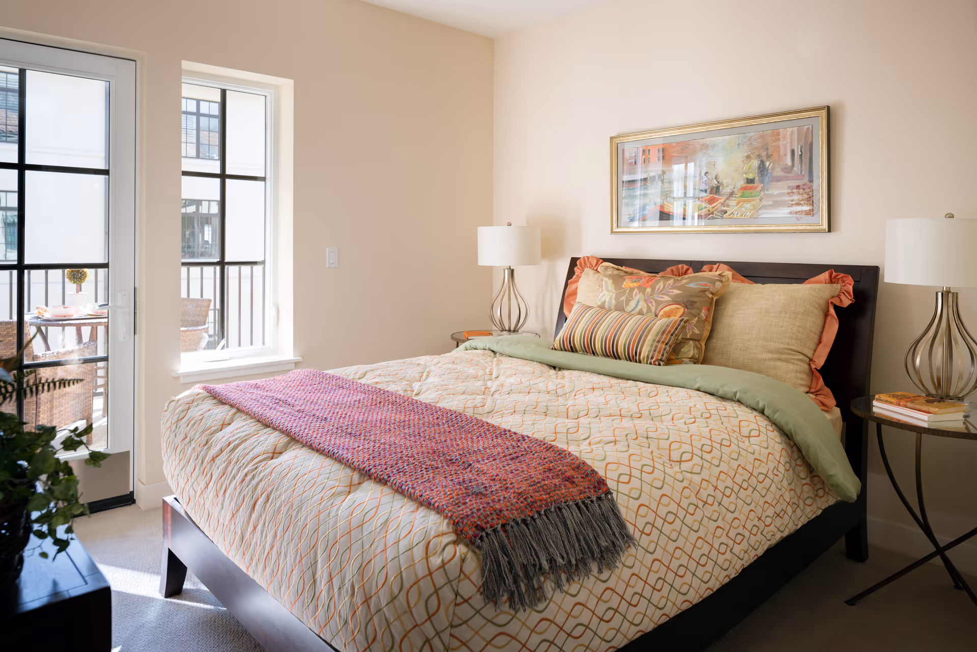 Sunlit bedroom with a neatly made bed topped with decorative pillows, bedside tables and lamps, and a door and window opening to a balcony.