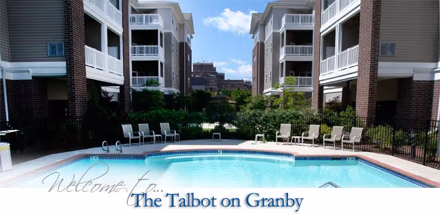 Outdoor swimming pool area between two multi-story residential buildings with lounge chairs arranged around the pool under a partly cloudy sky.