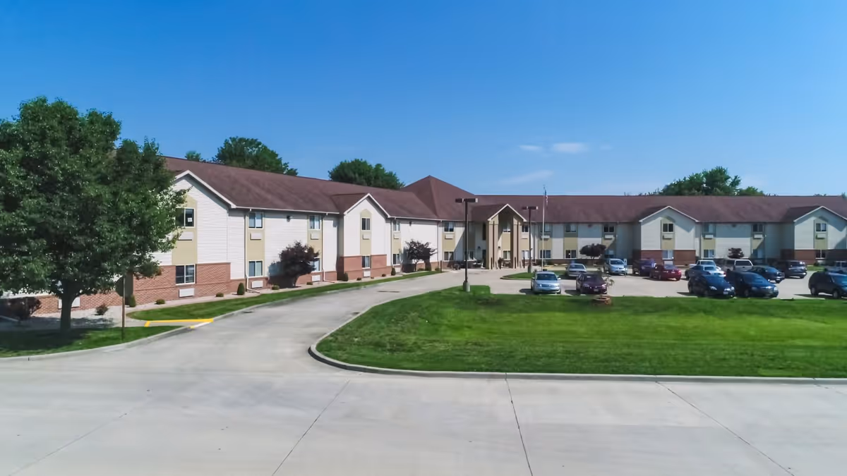 Front exterior of a low-rise senior living building with a curved driveway, lawn, and parked cars under a clear blue sky.