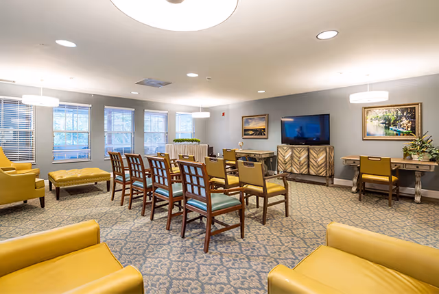A well-lit common area with multiple wooden chairs arranged in rows facing a large flat-screen TV mounted on the wall. The room features yellow armchairs, a patterned carpet, several windows with blinds, framed artwork on the walls, and two desks with chairs and decorative plants.
