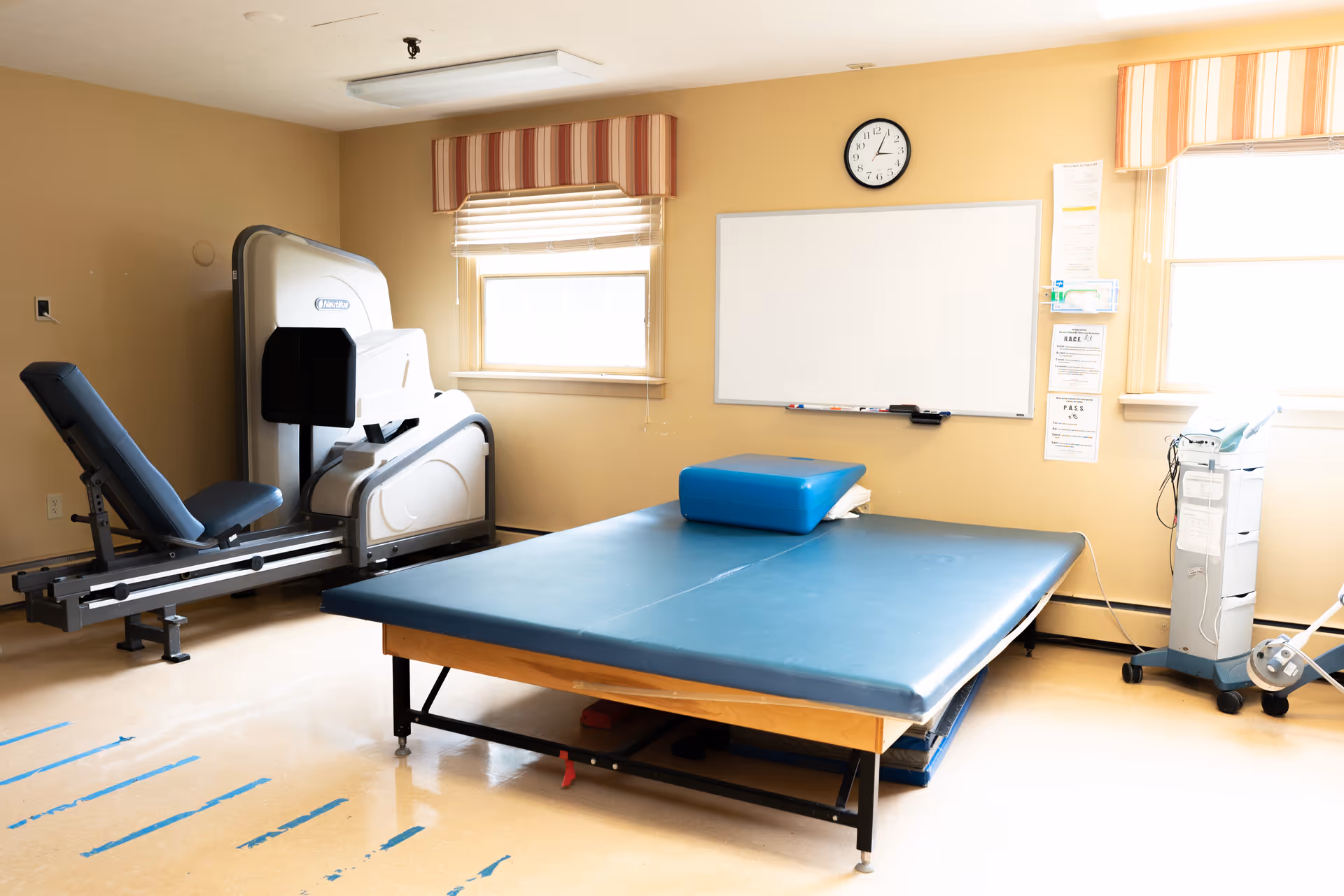 A physical therapy room with a large padded treatment table in the center, an exercise machine with a padded seat on the left, a whiteboard and clock on the wall, and two windows with striped valances allowing natural light into the room.