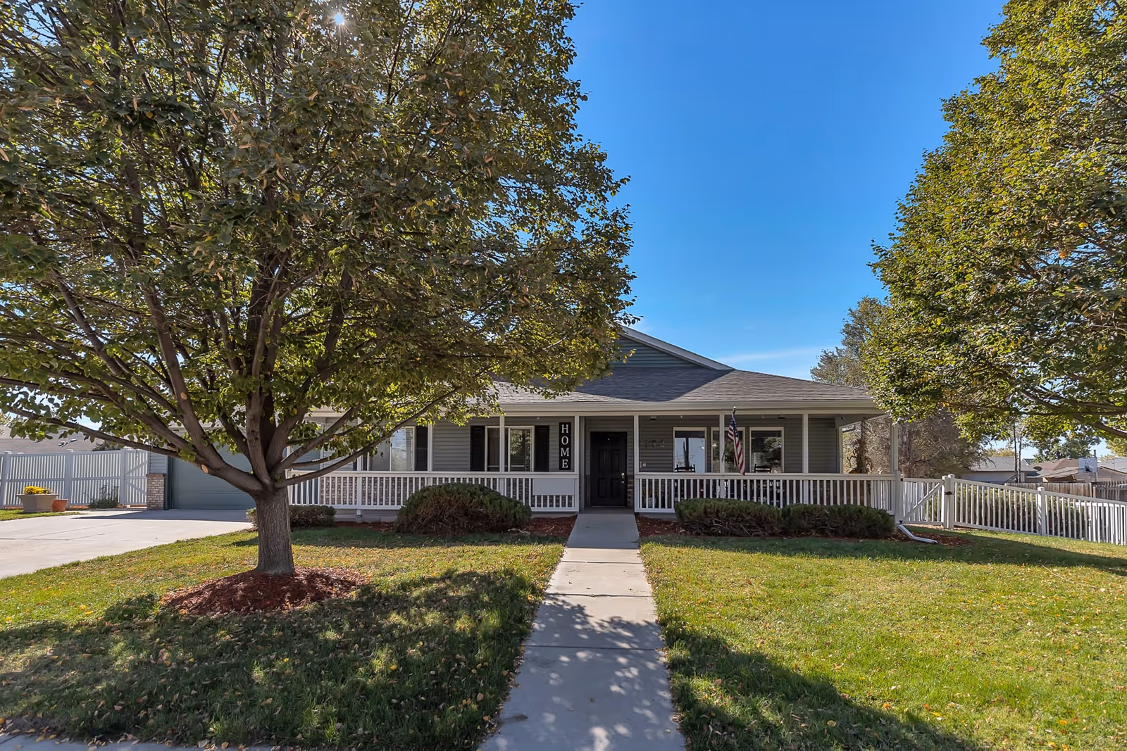 Front exterior view of a single-story house with a covered porch, white railing, and a black front door. There are two large trees on the lawn, a concrete walkway leading to the entrance, and an American flag hanging near the door. The sky is clear and blue.