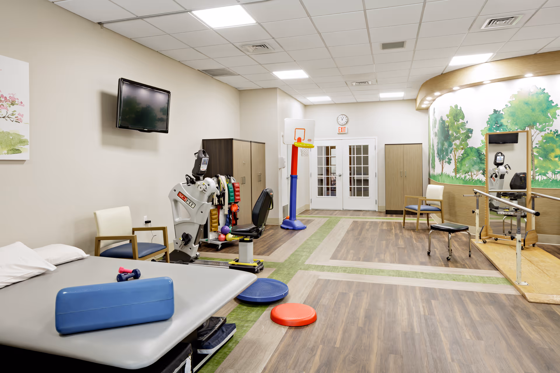 A physical therapy or rehabilitation room with exercise equipment including a recumbent bike, therapy table with weights and a bolster, chairs, a small basketball hoop, and a mirror with parallel bars. The room has wood-patterned flooring with green accents and a mural of trees on one wall.