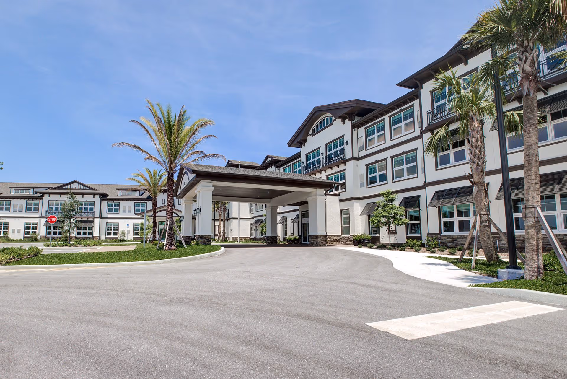 Front exterior of a multi-story senior living building with a covered entrance, palm trees, and a curved driveway.