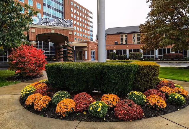 Exterior view of St. Ann's Community building with a landscaped garden featuring colorful flowers and neatly trimmed bushes in the foreground. The building has a brick facade with large windows and a covered entrance.