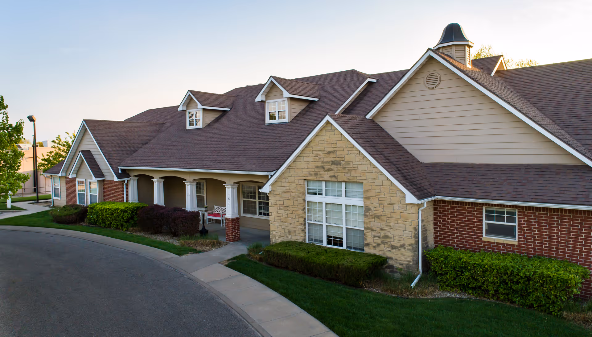 Exterior view of a single-story assisted living facility building with a combination of brick and stone facade, multiple windows, a covered porch with white columns, and a well-maintained lawn and shrubs along a curved driveway.