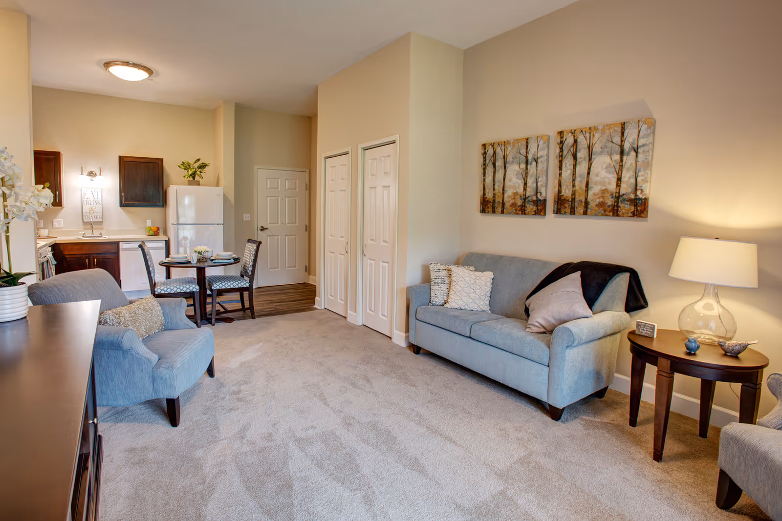A cozy living area with a light blue sofa adorned with pillows and a dark throw blanket, a matching armchair, a wooden side table with a lamp and decorative items, and two paintings of trees on the wall. In the background, there is a small dining table with two chairs set with plates and cups, and a compact kitchen area with a refrigerator, dishwasher, and cabinets. The room has beige walls and carpeted flooring.
