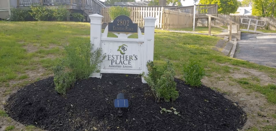 Outdoor view of a sign for Esther's Place Assisted Living at 2802, surrounded by a mulched garden bed with small green shrubs, with a building and ramp in the background.