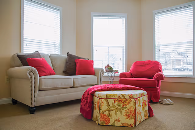 Bright living room with a beige sofa, red armchair, floral ottoman and three large windows.