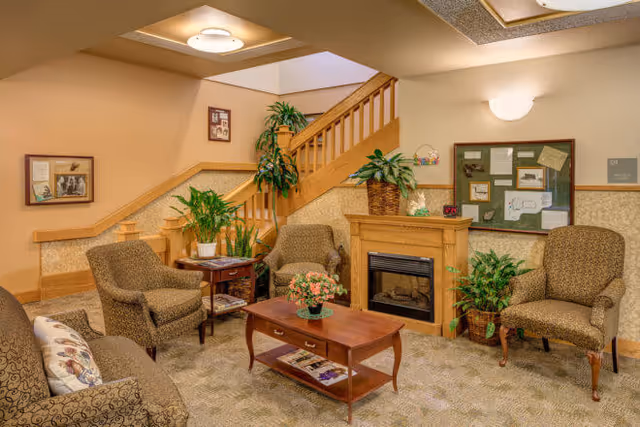 A cozy living room area in Marlow Manor Assisted Living Facility featuring four upholstered armchairs arranged around a wooden coffee table with a floral centerpiece. There is a wooden fireplace with a plant on top, a bulletin board on the wall, and several potted plants placed around the room. A staircase with wooden railings is visible in the background, along with framed pictures on the walls.