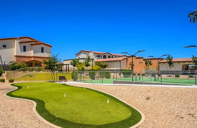 Outdoor courtyard at Fellowship Square Surprise featuring a putting green, fenced sports courts, and low-rise Mediterranean-style buildings under a clear blue sky.
