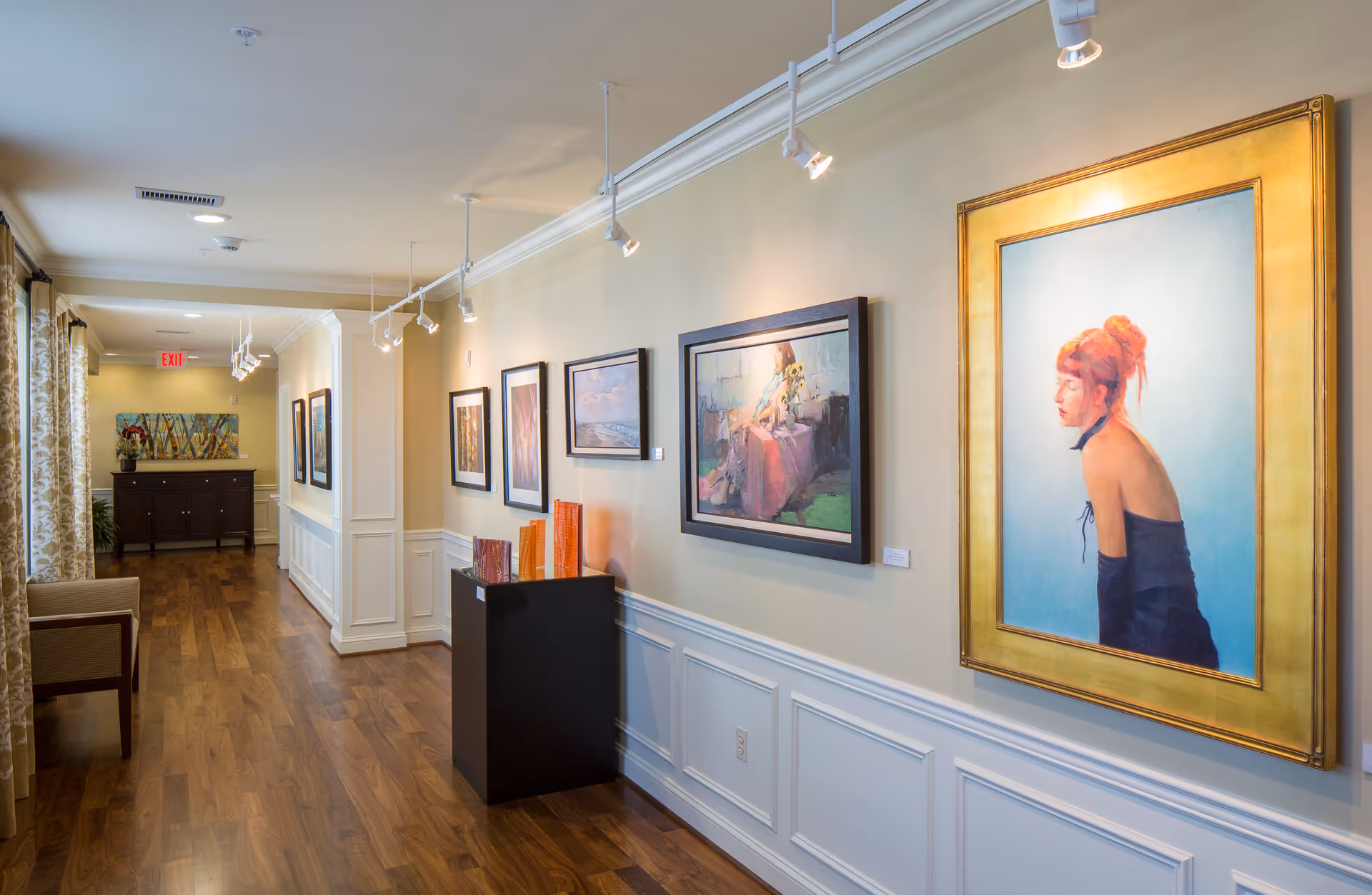 A well-lit hallway in a senior living facility with wooden flooring and cream-colored walls adorned with framed paintings and artwork. There is a black pedestal displaying decorative items, a chair near a window with patterned curtains, and a cabinet with a plant and artwork at the end of the hallway.