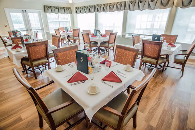 A bright dining room with multiple square tables covered with white tablecloths, each set with red folded napkins, white cups, saucers, silverware, and menus. The room has large windows with sheer white curtains and valances, and wooden flooring.