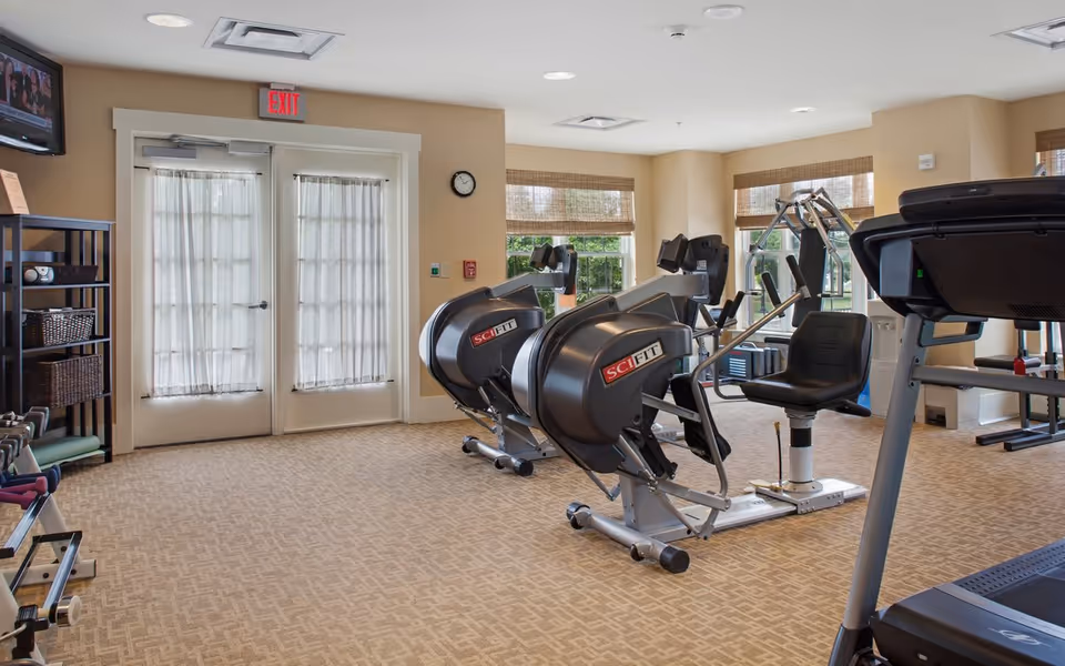 Bright fitness room with cardio machines (SciFit recumbent/rowing machines), a treadmill, weight shelves, windows, and double doors.