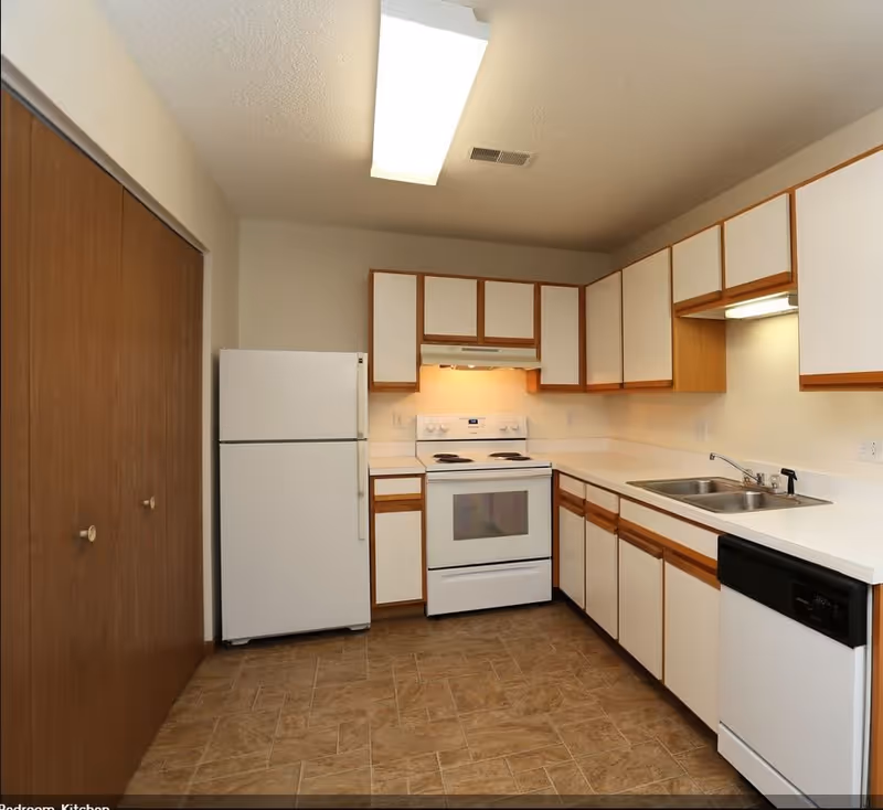 A clean kitchen with white cabinets featuring wooden trim, a white refrigerator, an electric stove with an oven, a double sink, and a dishwasher. The floor has a brown tile pattern, and the ceiling has a rectangular fluorescent light fixture.