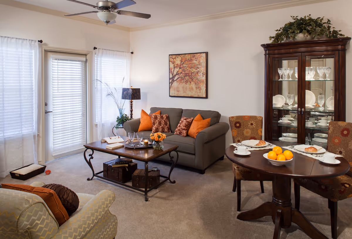 A cozy living room with a gray sofa adorned with orange and patterned cushions, a wooden coffee table with books, glasses, and a flower vase, a round dining table set with plates, cups, and a bowl of oranges, a glass cabinet displaying glassware and dishes, an armchair with cushions, and a ceiling fan above. Light filters through sheer white curtains covering windows and a door.